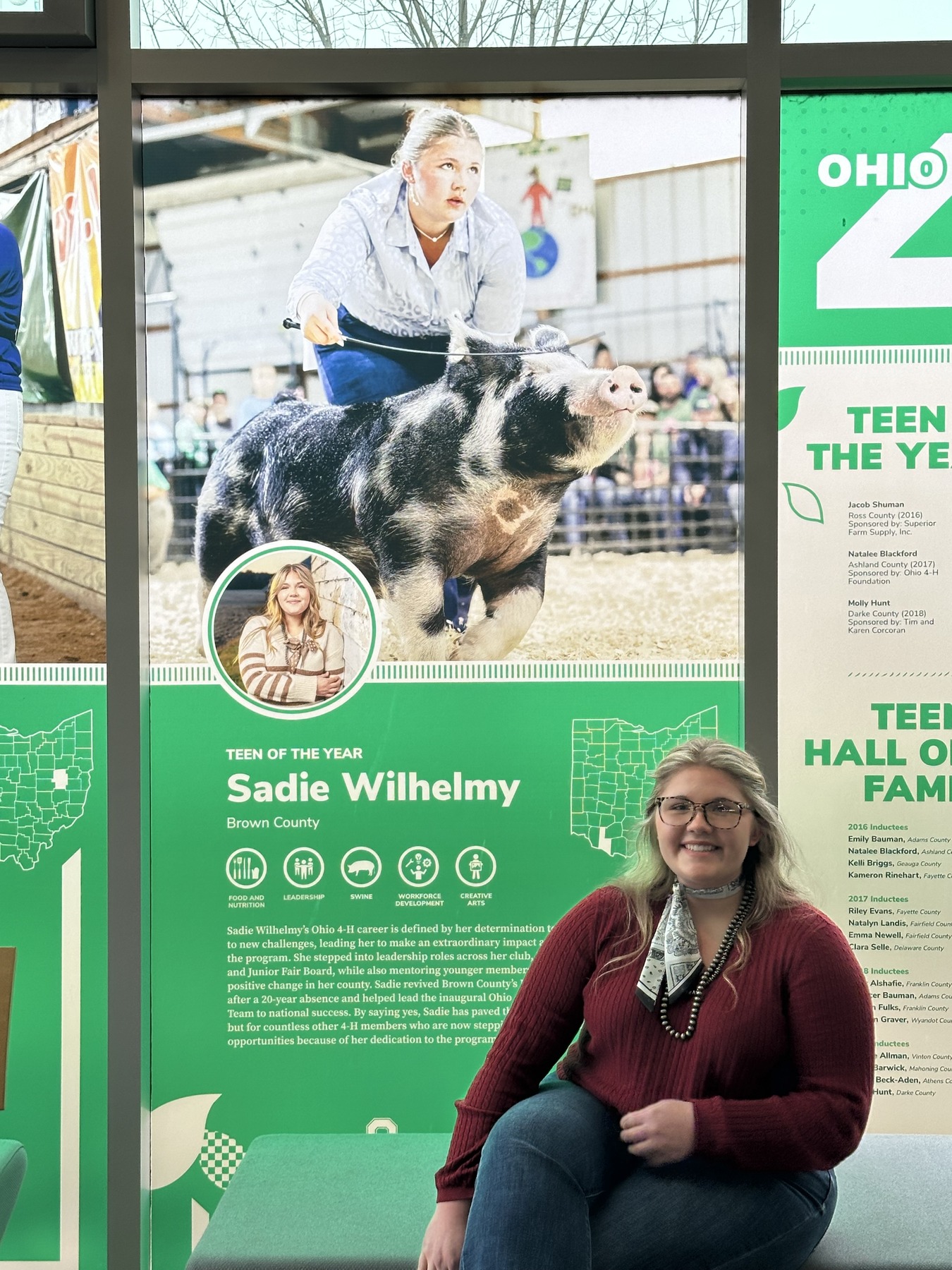 Sadie Wilhelmy seated in front of the State of Ohio 4-H Teen of the Year display board